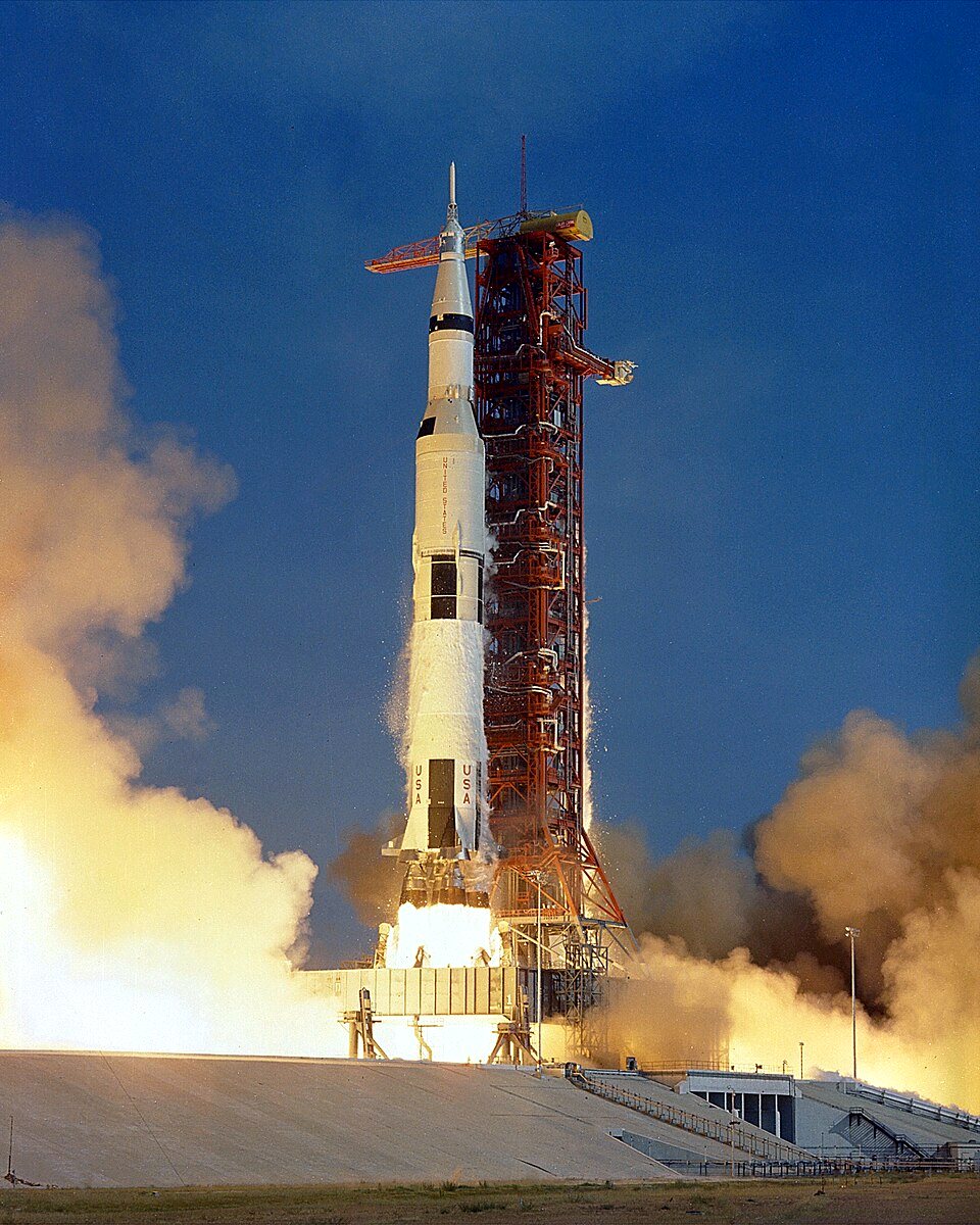 A tall white and black Saturn V rocket on its pad rises on a column of fire and smoke against a deep blue sky.