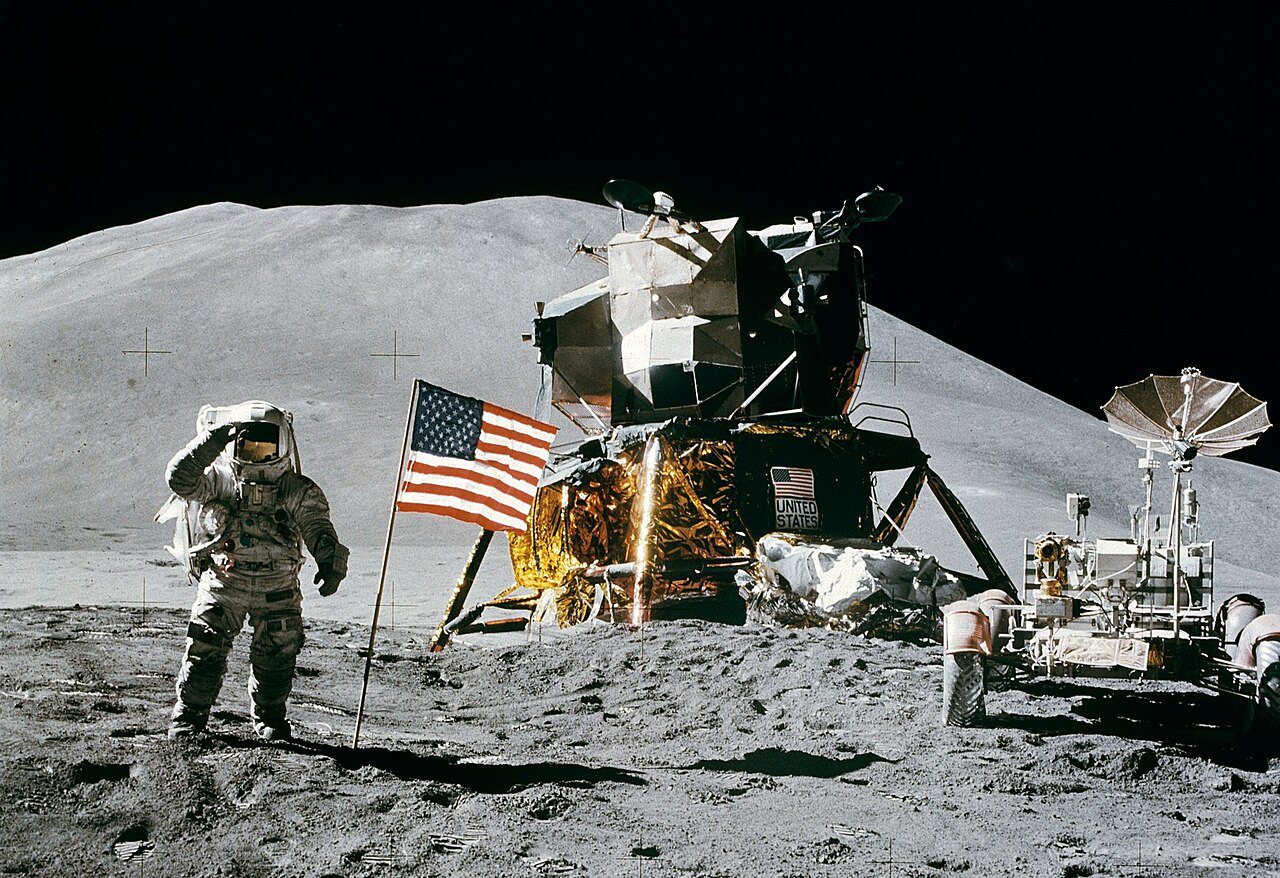 An astronaut in a white spacesuit salutes beside an American flag, with a lunar module, a rover and a mountain behind him.
