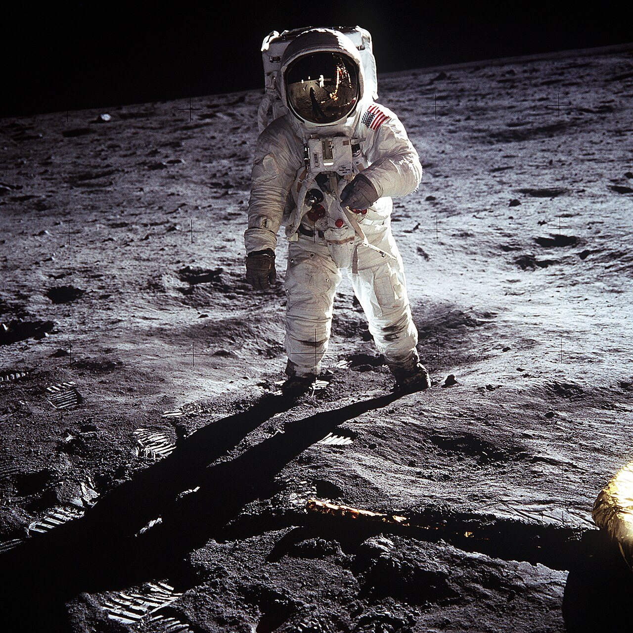 Astronaut Buzz Aldrin in a white spacesuit and gold visor stands on gray lunar soil, with a long shadow stretching across the ground.