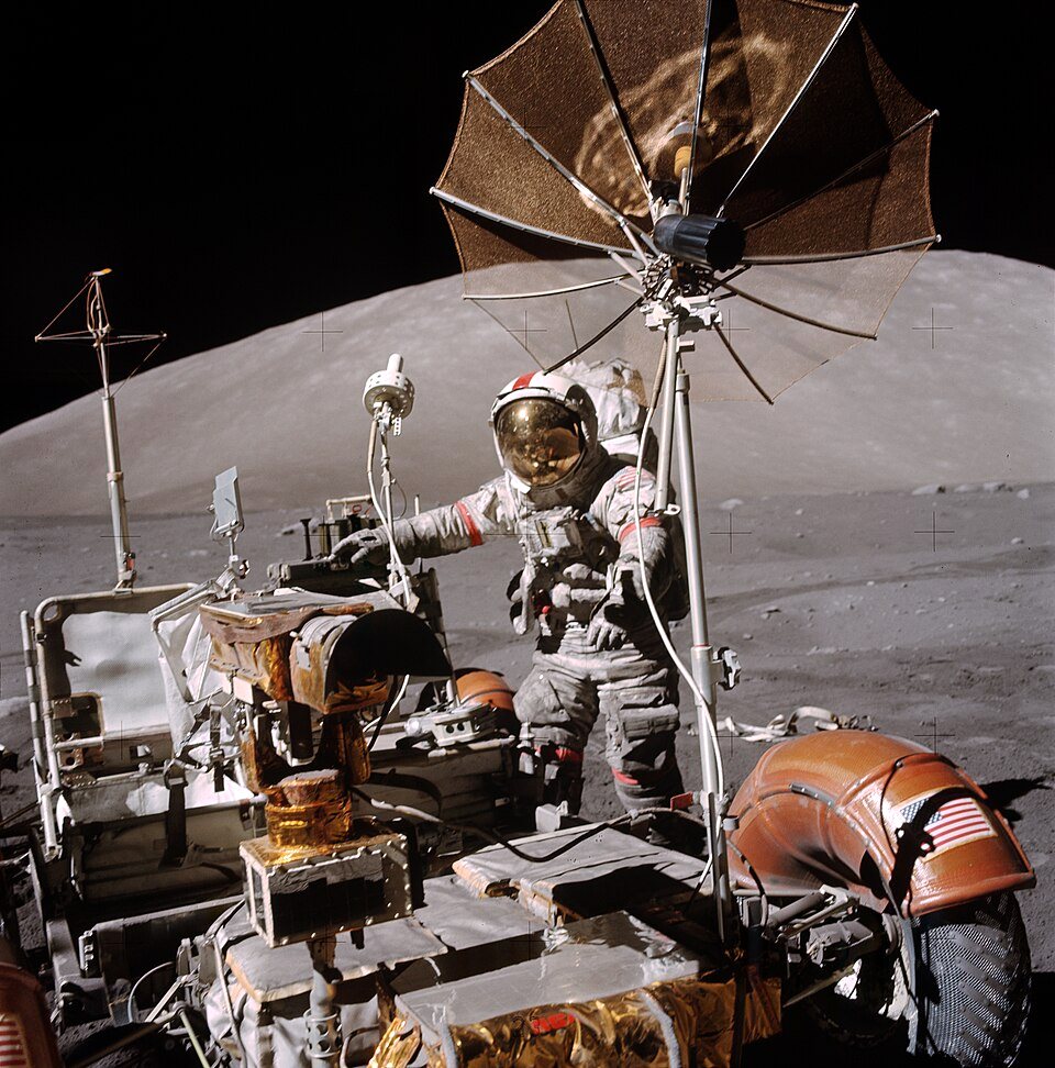An astronaut in a gold-visored helmet stands behind a lunar rover loaded with equipment, its mesh dish open above and a gray hillside behind.