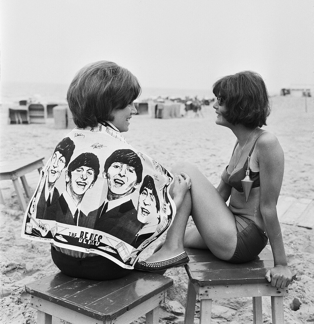 Two young women sit on wooden stools on a beach; one wears a bath towel printed with four drawn faces of the Beatles over her back, and the other, in a swimsuit, looks at her.