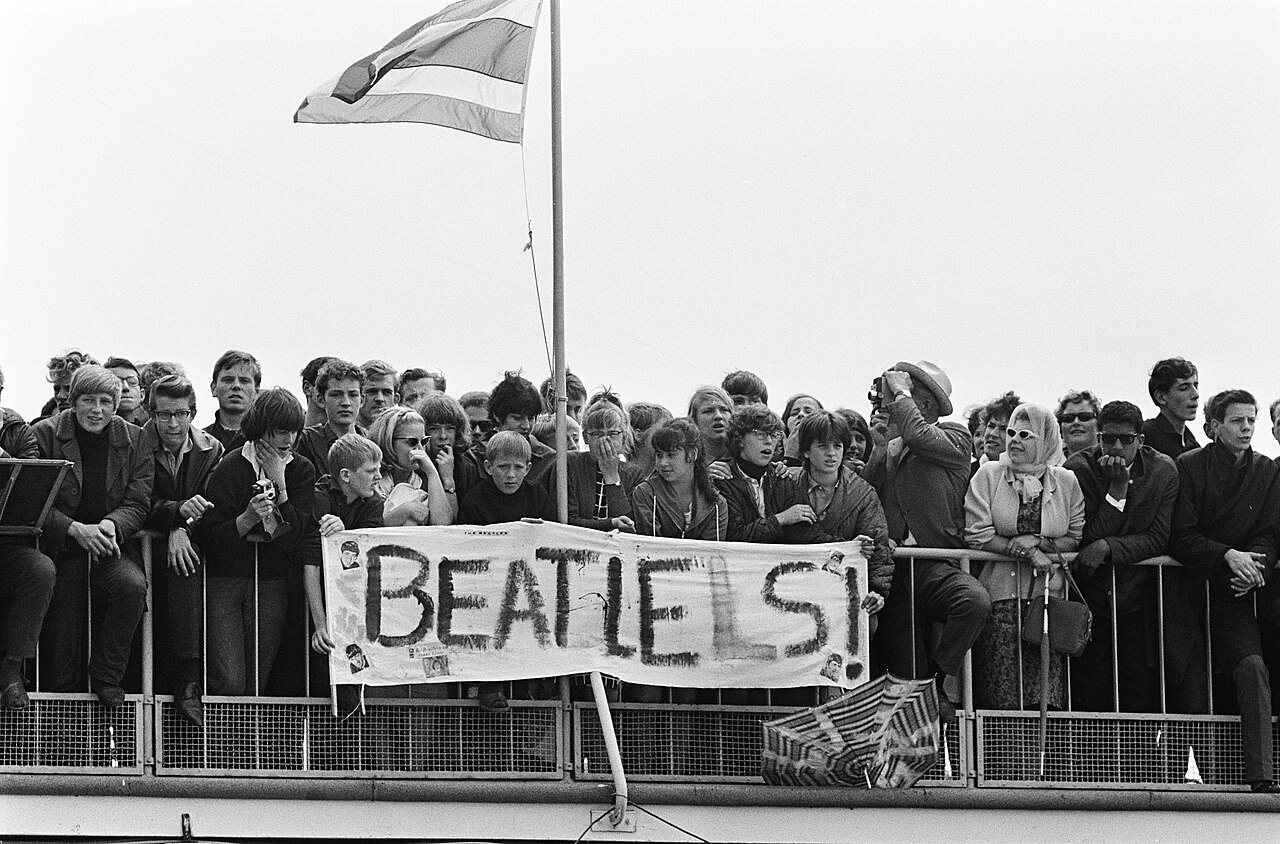 A crowd of young and older people packs a mesh railing behind a hand-lettered banner that reads Beatles, with a striped flag on a pole above them and a folded umbrella at the foot of the banner.