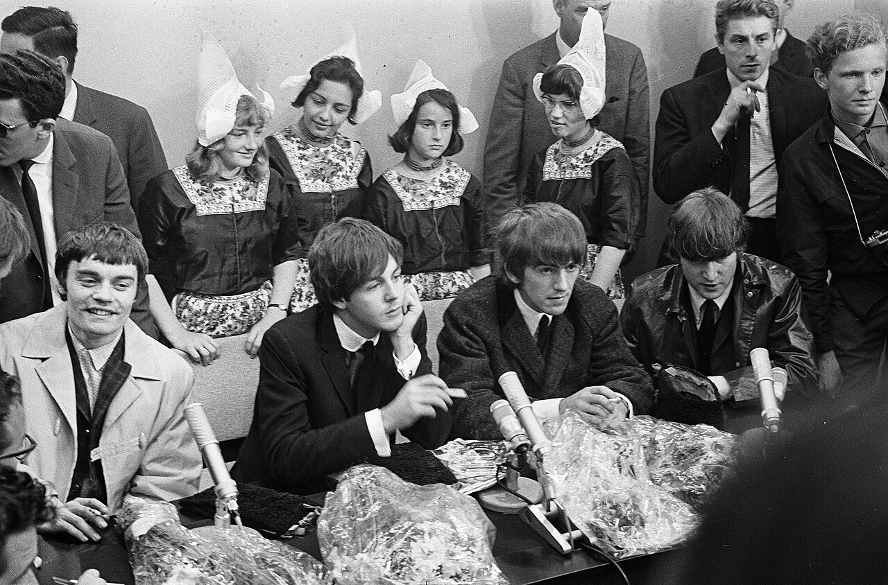 Four men sit at a table crowded with microphones and cellophane-wrapped bouquets, with four girls in Volendam costume and white caps seated in a row behind them and other people standing around.