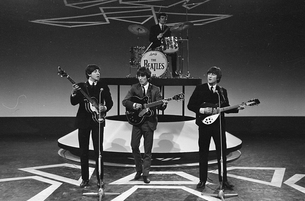 Three men with guitars stand on a round stage with a zigzag floor pattern, and a drummer sits behind a kit with a drum head reading The Beatles.