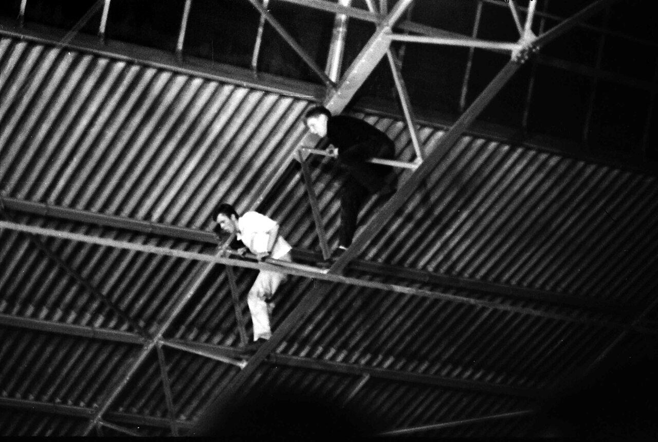Two small figures balance on steel roof trusses under a dark corrugated roof, one of them in a white shirt, high above the floor of a hall.