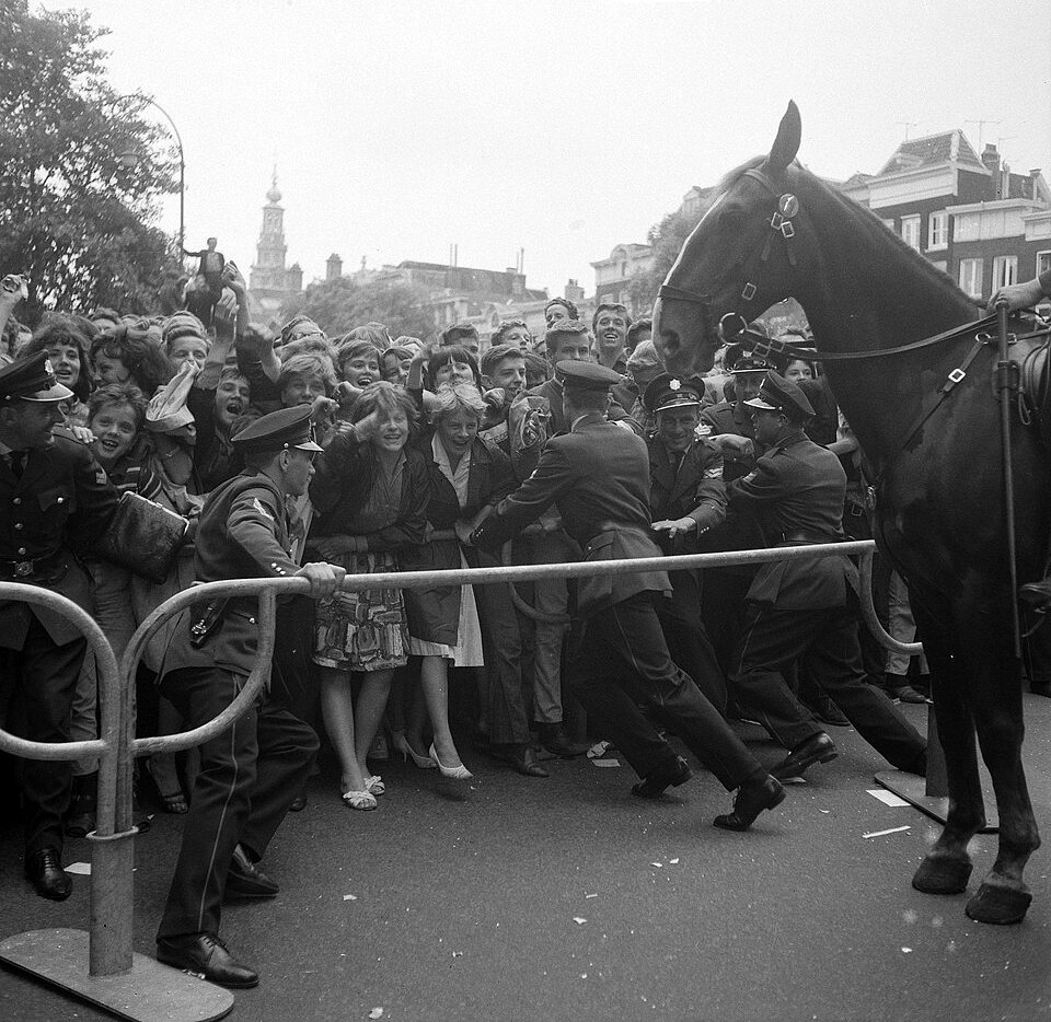 Uniformed police lean into a steel barrier holding back a crowd of young people, with a tall police horse at the right edge and a tower in the distance.