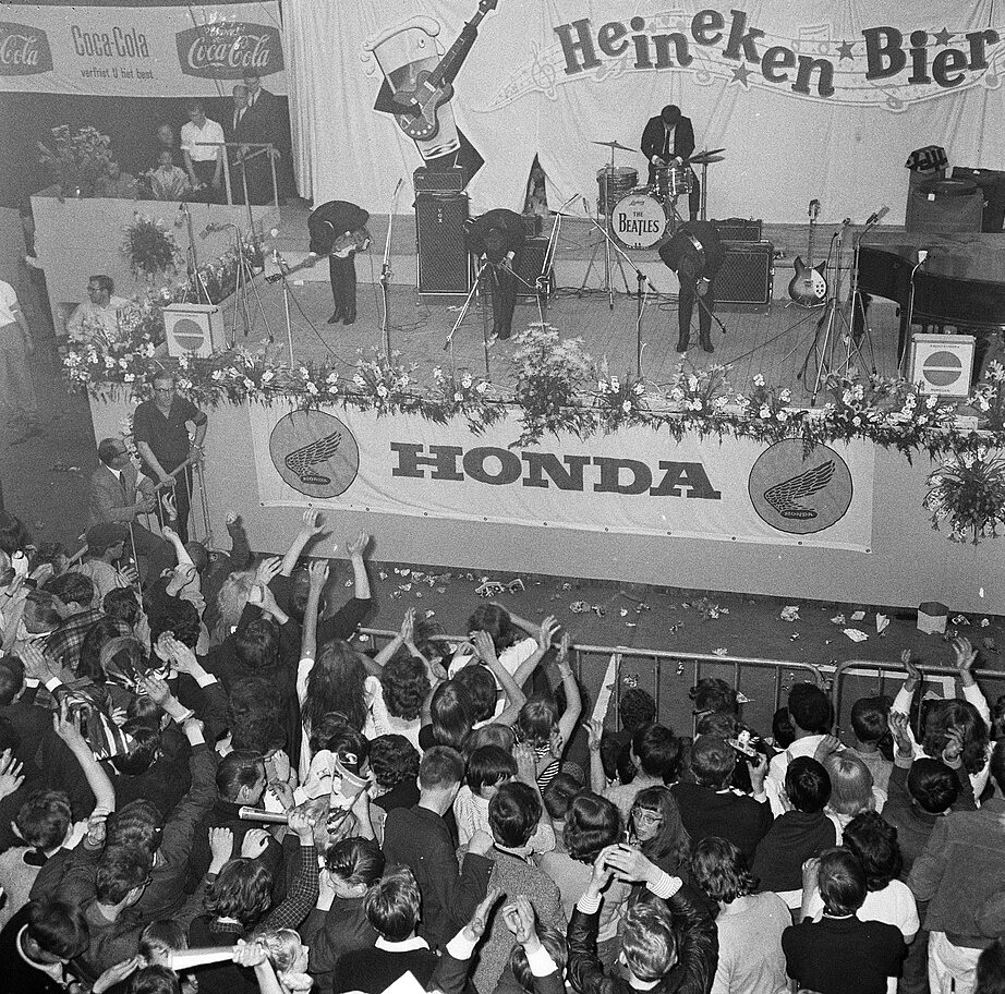 Seen from above, a stage with a Heineken Bier backdrop and a Honda banner, three guitarists bowing in front of a drum kit marked The Beatles, and a crowd with raised arms pressed against a barrier below.