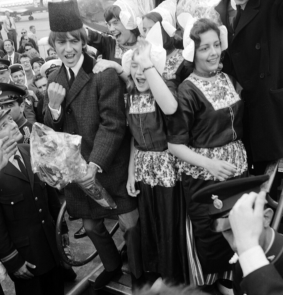 On an airport staircase, a young man in a tall dark cap grins and raises a fist, holding a bouquet, while two girls in Volendam costume and white caps laugh beside him and uniformed officers stand below.