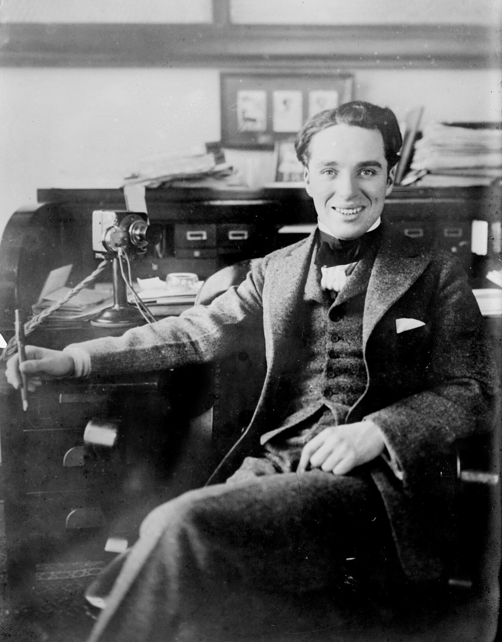 A young man with dark wavy hair, a vest, a bow tie and a loose jacket sits at a desk with his arm resting on it, smiling at the camera.
