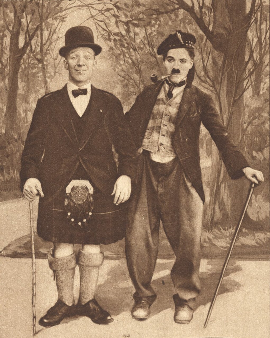 Two men in costume pose in front of a painted woodland backdrop, one in a bowler hat, kilt and long socks, the other in a small hat, mustache, baggy trousers and a tight jacket, each holding a cane.