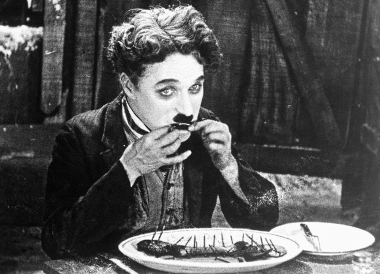 Chaplin, in a dark jacket, raises a bootlace to his mouth over a plate holding a boiled shoe, with a fork and a second plate beside it.