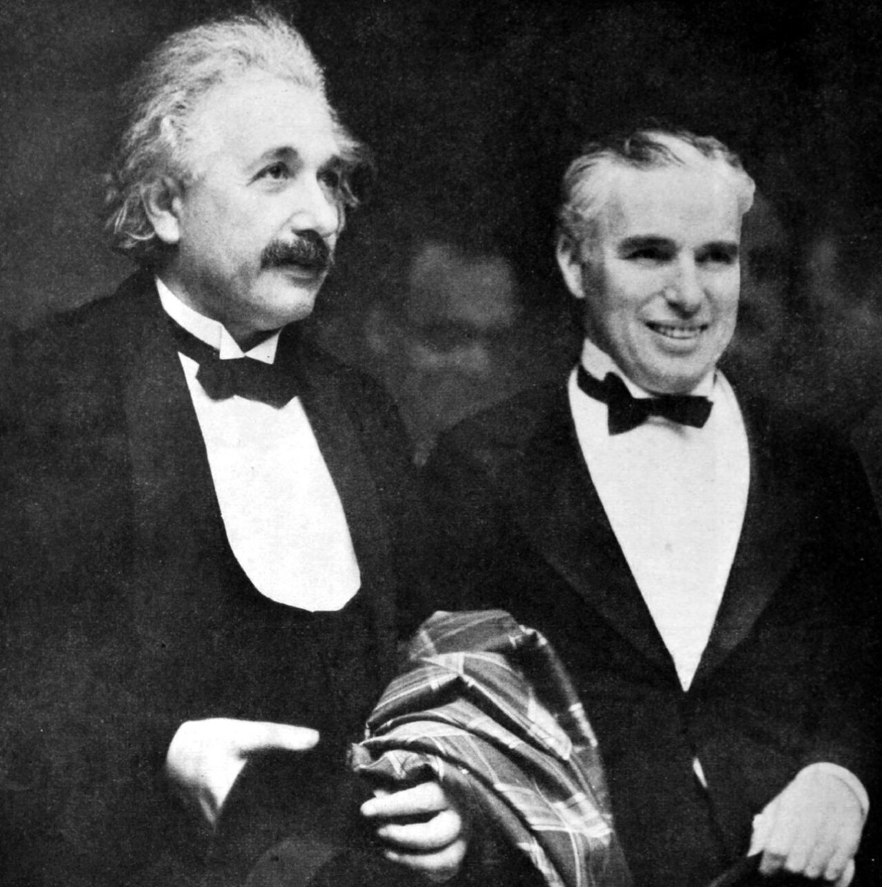 Albert Einstein, with tousled gray hair and a mustache, and Charlie Chaplin, smiling, sit side by side in dinner jackets and bow ties, with a plaid wrap over Einstein’s arm.