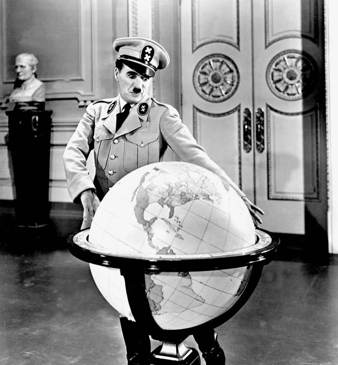 A man in a military uniform with a peaked cap and a toothbrush mustache stands behind a large globe on a stand, resting his hands on it, in front of tall double doors.