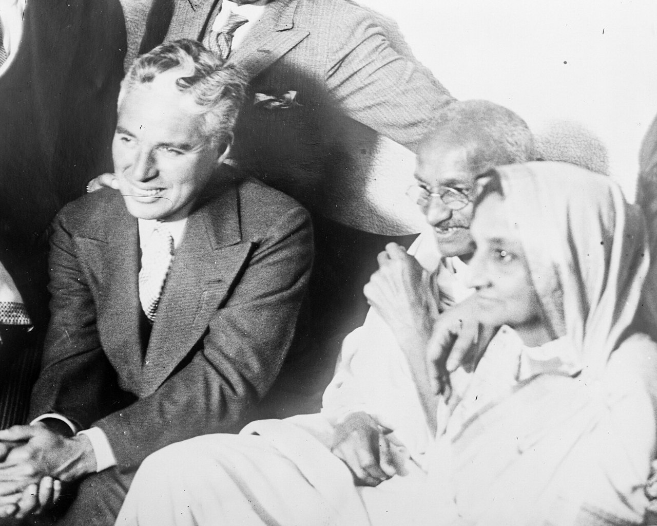 Chaplin, in a suit, sits smiling with his hands clasped beside Mahatma Gandhi, who wears glasses and a white shawl, with a woman in a light head covering seated in front of them.