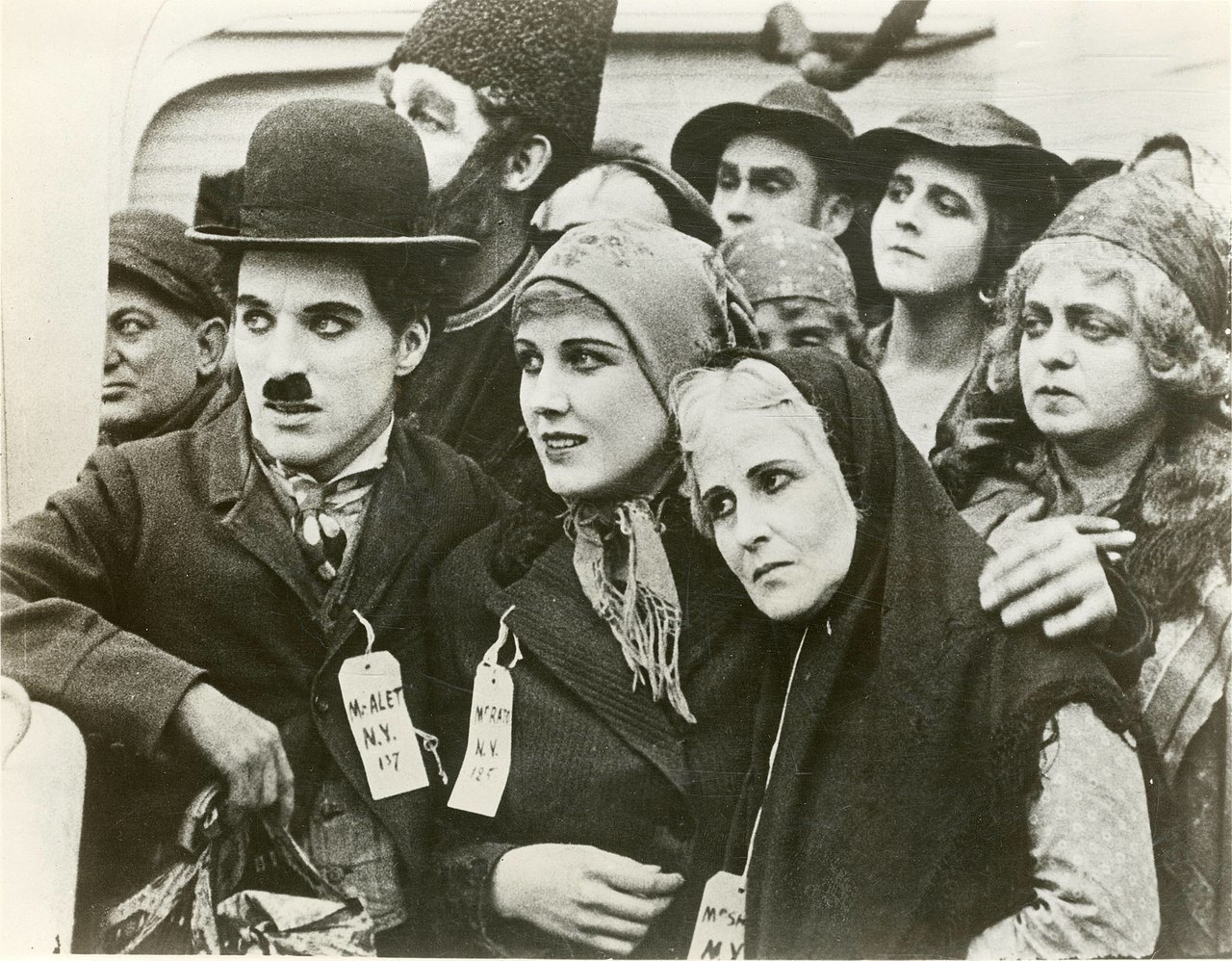 Chaplin in a bowler hat and toothbrush mustache stands shoulder to shoulder with weary passengers in shawls and caps, a paper tag pinned to his coat, while a woman beside him looks worried.