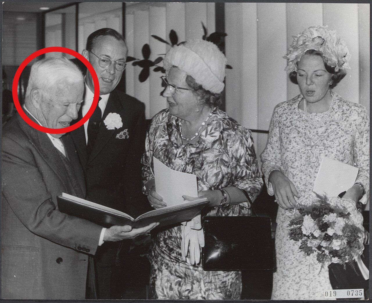 An older man with white hair, circled in red, holds an open book on the left, while a tall man in a dark suit, a woman in a white hat and a younger woman holding flowers stand beside him.