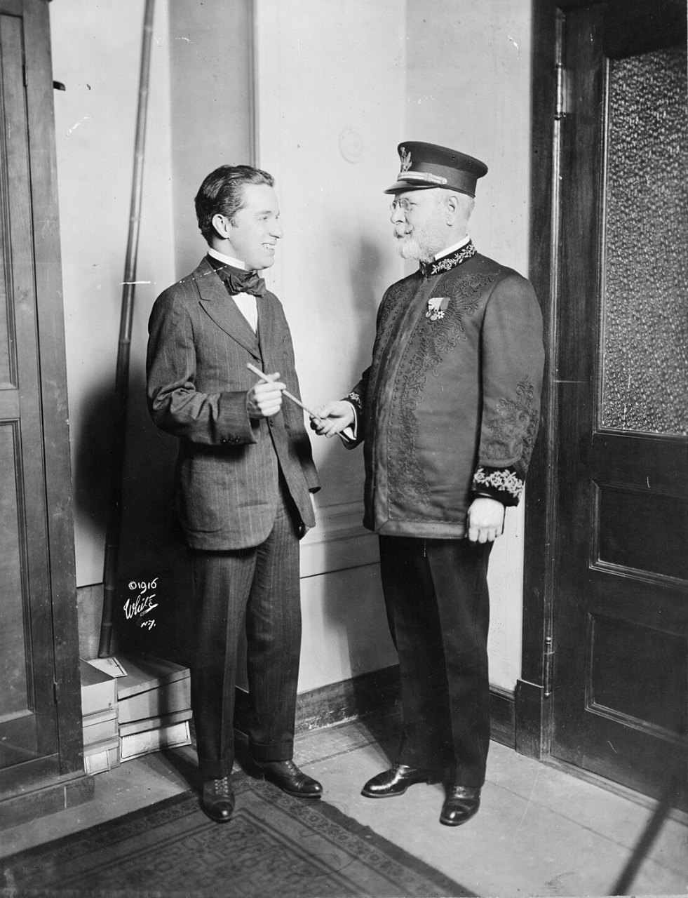 A man in a dark suit and bow tie holds a small object in one hand while talking with an older man in a band uniform and peaked cap, in a corridor with wooden doors.