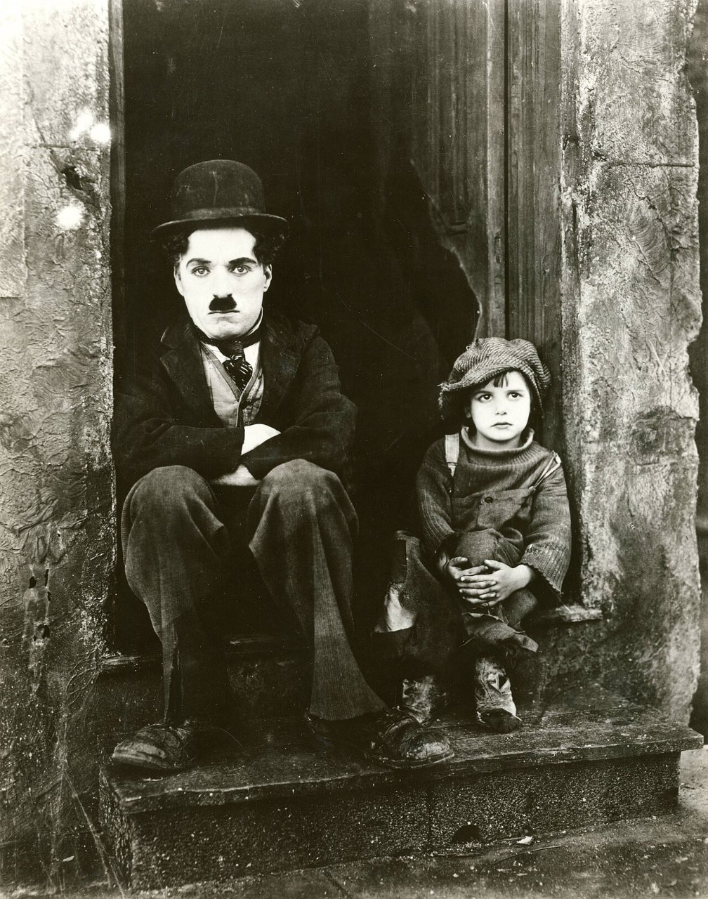 A man in a bowler hat, small mustache and shabby suit sits on a stone doorstep next to a small boy in an oversized cap and ragged clothes, both looking at the camera.