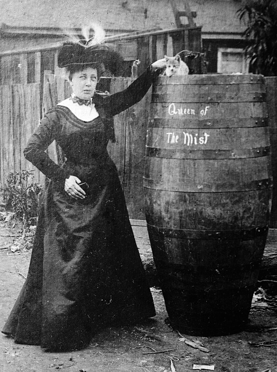 A woman in a long dark dress and a feathered hat stands beside a tall wooden barrel lettered Queen of the Mist, one arm raised toward a small cat sitting on top.