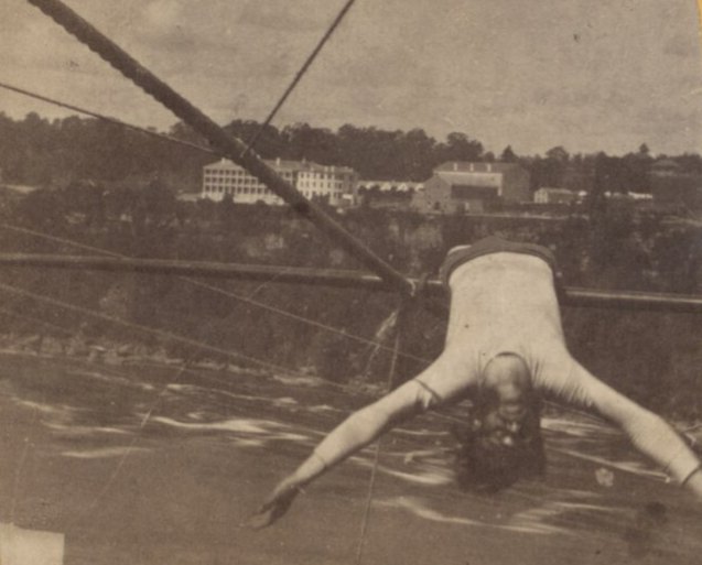 A man in a pale sleeveless shirt hangs upside down from a rope above a river with his arms spread wide, with cables and distant buildings behind him, in one view from an old stereo card.