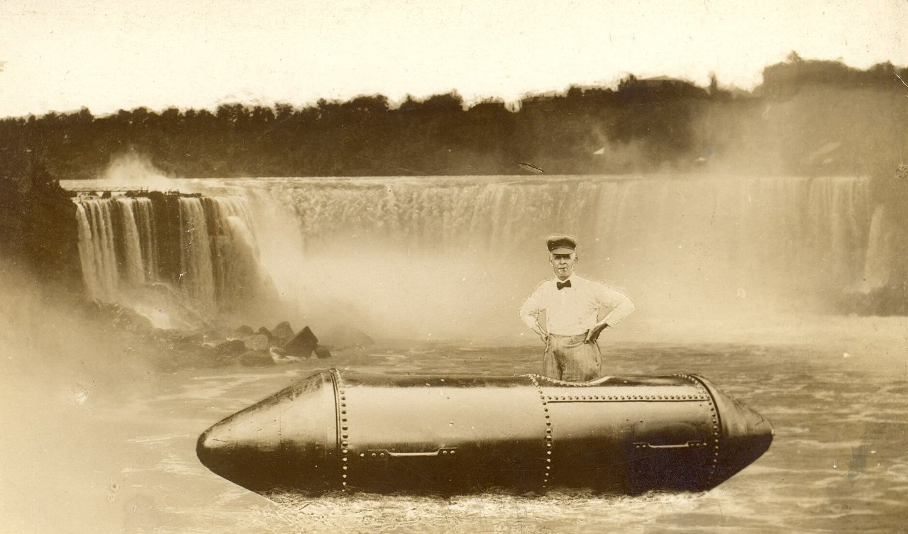 A man in a white shirt, bow tie and cap stands with his hands on his hips in the open top of a long, riveted steel barrel floating on the water, with Niagara’s Horseshoe Falls and mist behind him.