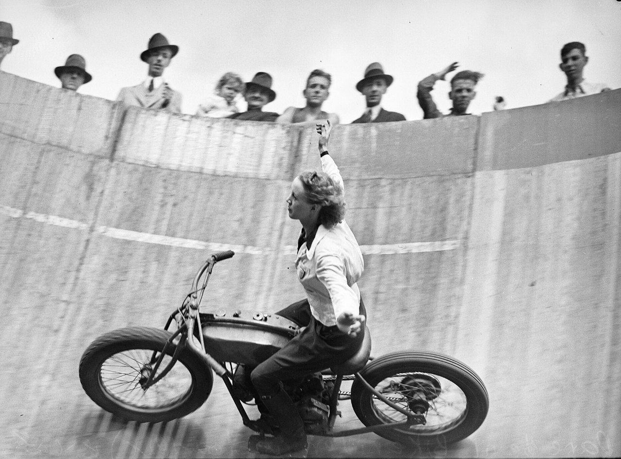 A woman in a white blouse rides a motorcycle along the steeply banked wall of a circular wooden track, one arm raised, while a row of men in hats and a small child watch from the rim above.
