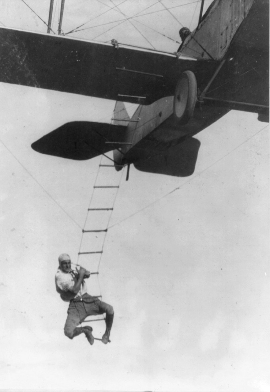 A man in a light shirt and dark trousers hangs from a rope ladder beneath a biplane high above the ground, gripping the rungs with his arms and legs against a pale sky.
