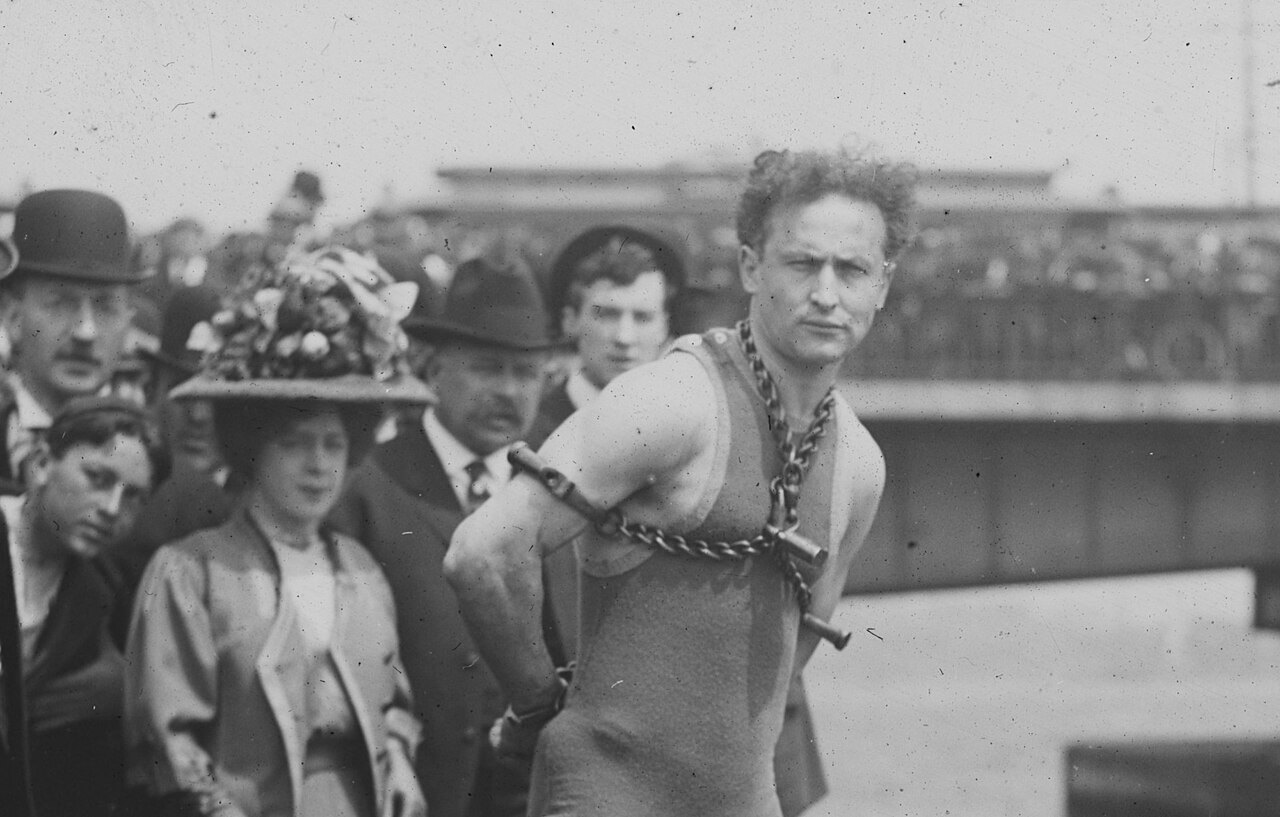 A man in a sleeveless knit swimsuit stands with a heavy chain wound around his neck and chest and his arms held behind him, beside a woman in a large flowered hat and several men in hats, with a bridge and a crowd behind them.