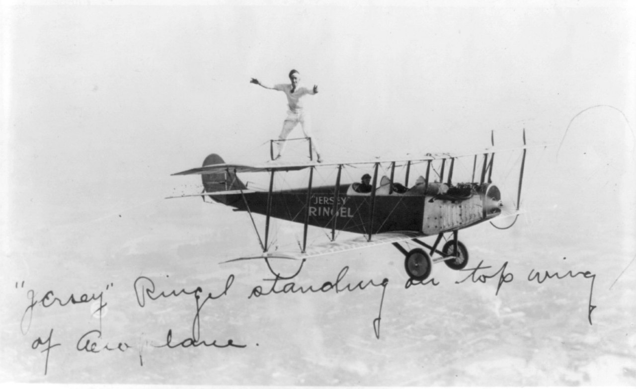 A man stands upright with his arms out on the top wing of a biplane flying high above the clouds, the plane’s side marked with his name, and a line of handwriting runs across the bottom of the print.