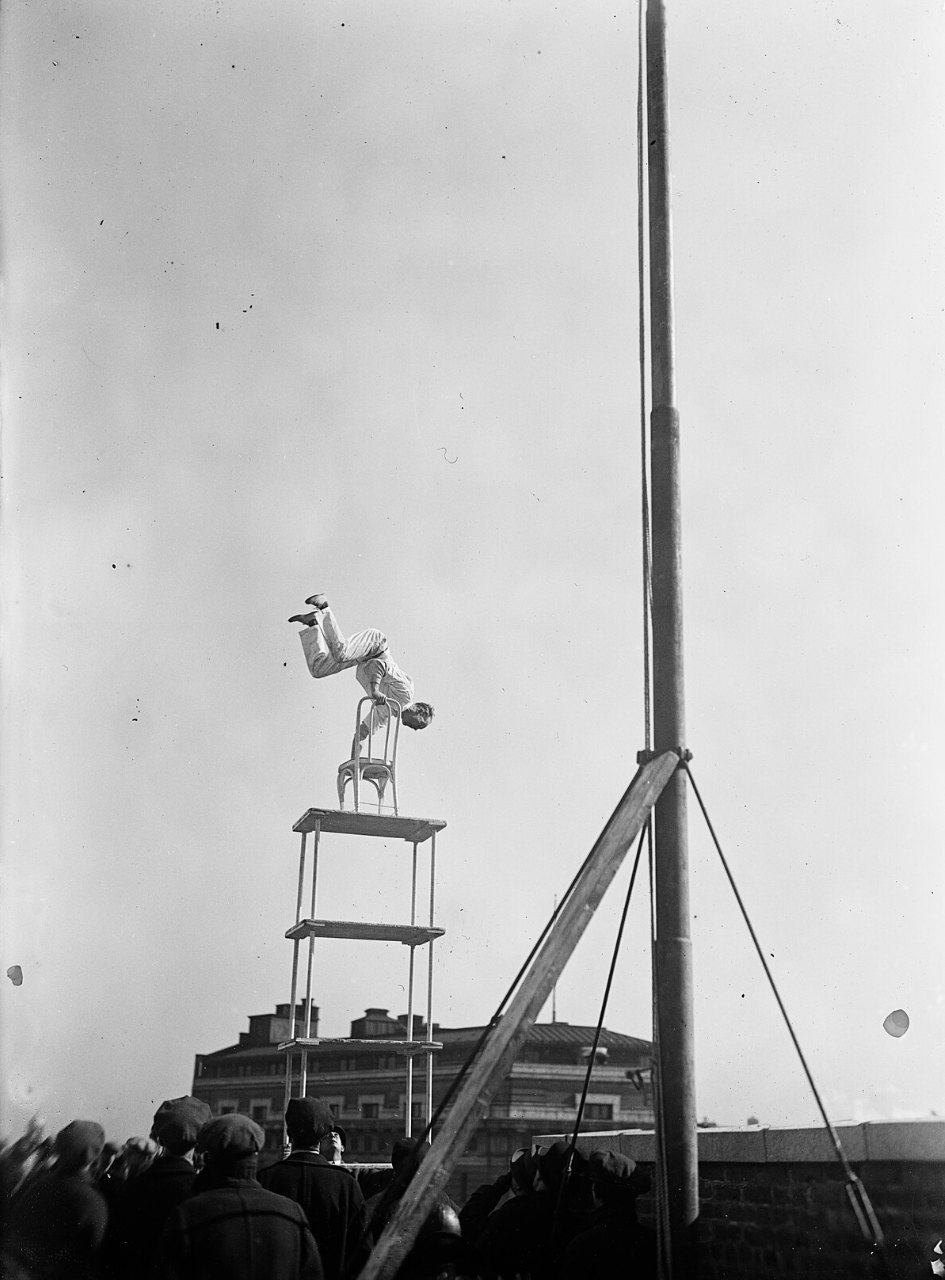 A man does a handstand on a chair set on top of a tall open frame, beside a pole braced with a timber and guy wires, while a crowd in caps watches from below.