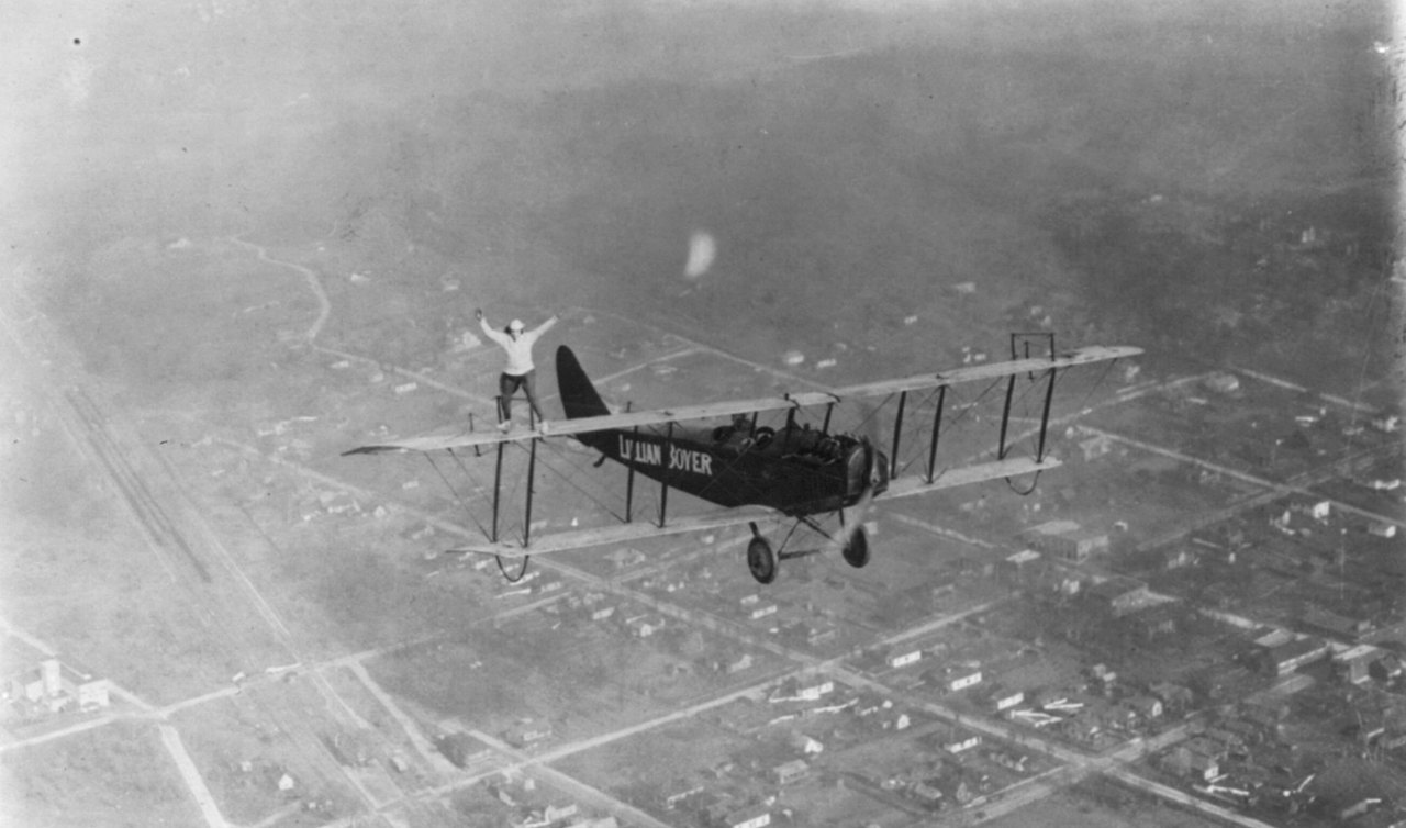 A woman stands on the top wing of a biplane with her arms stretched out, high above a city laid out in a grid of streets.