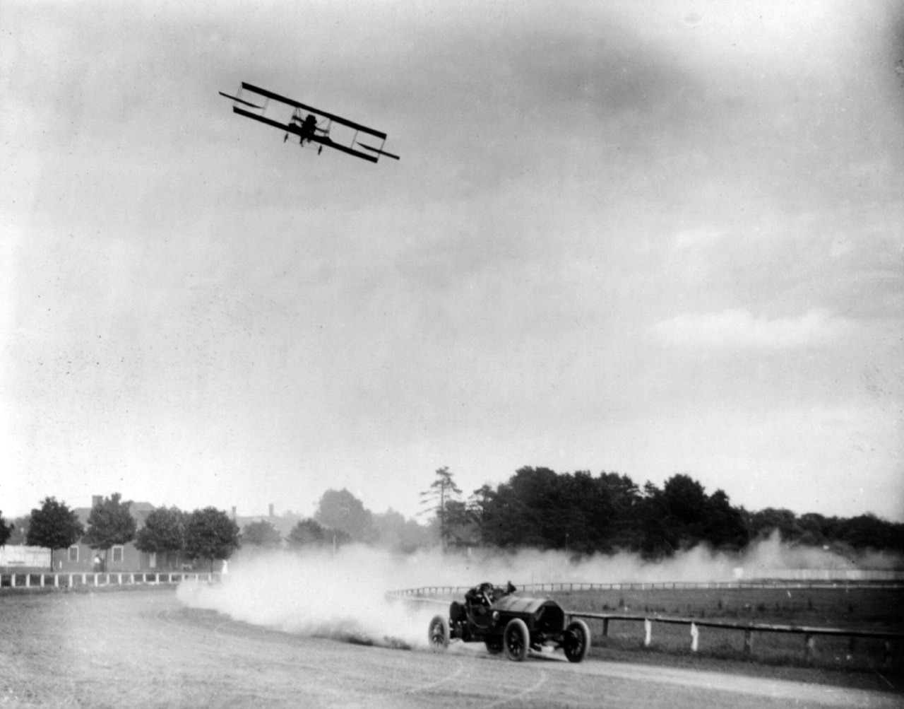 A race car speeds along a dirt track trailing a cloud of dust, while a biplane flies above it in a gray sky, with trees and a fence in the distance.