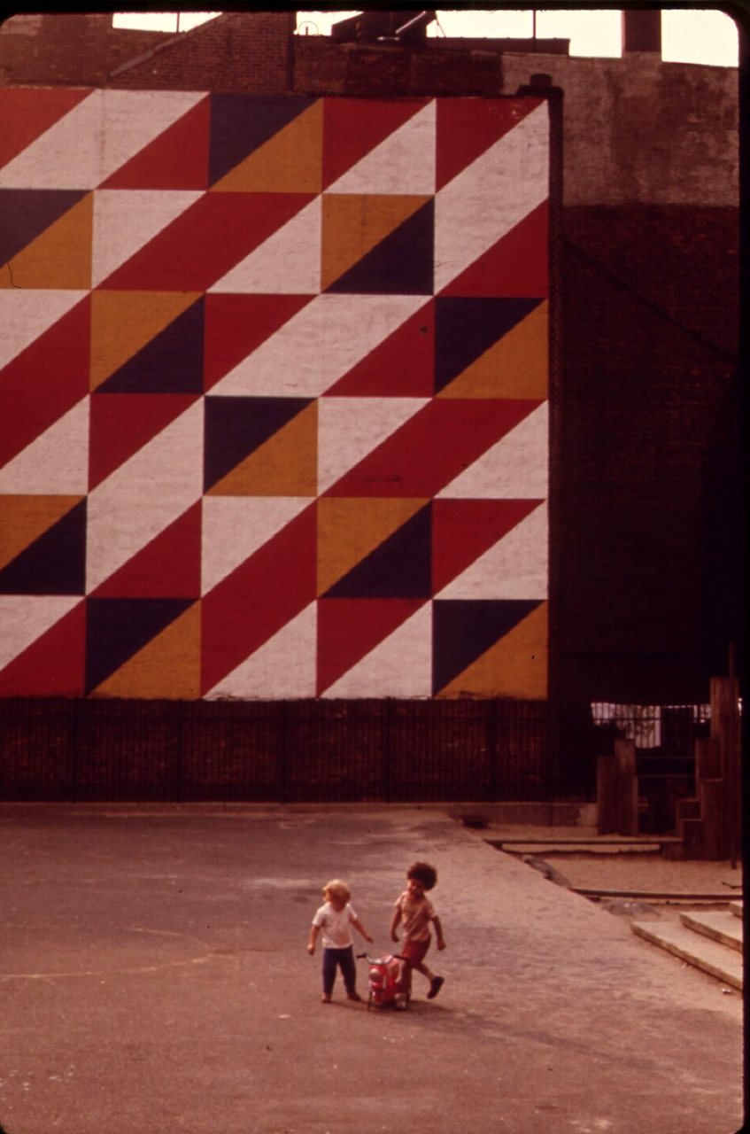 A mural of red, orange, navy and white triangles on a wall, with two small children walking in the paved park below.