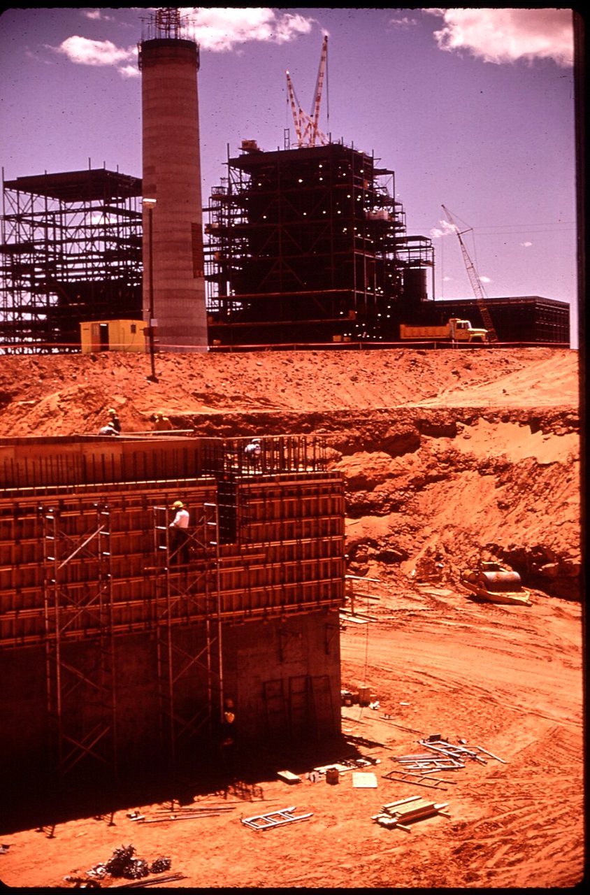 A power plant under construction in red desert soil, with scaffolding, formwork and a tall concrete chimney.
