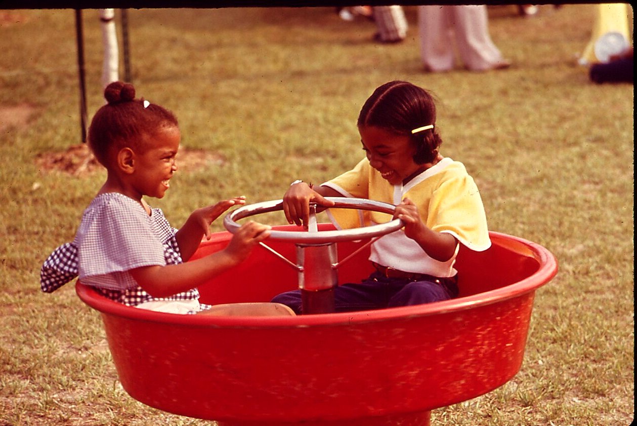 Two smiling girls sit in a round red spinning ride with a steering wheel in a grassy park.