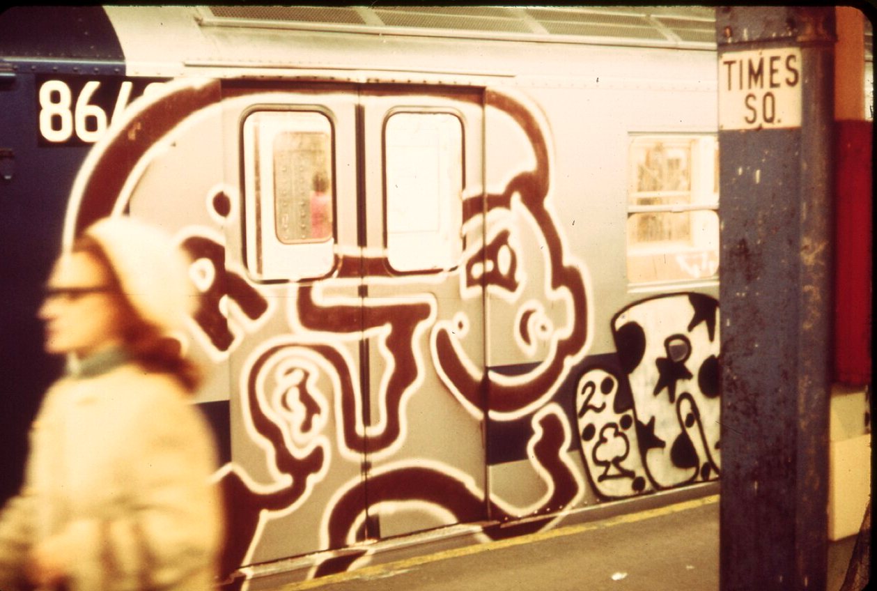 A subway car painted with a giant outlined face stands at the Times Square station while a blurred commuter walks by.