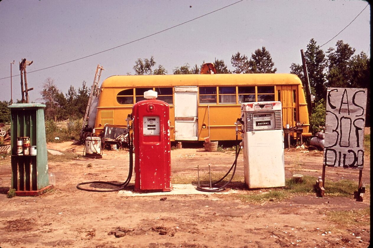 Three gas pumps and a hand-painted sign on a dirt lot, with an orange bus parked behind them.