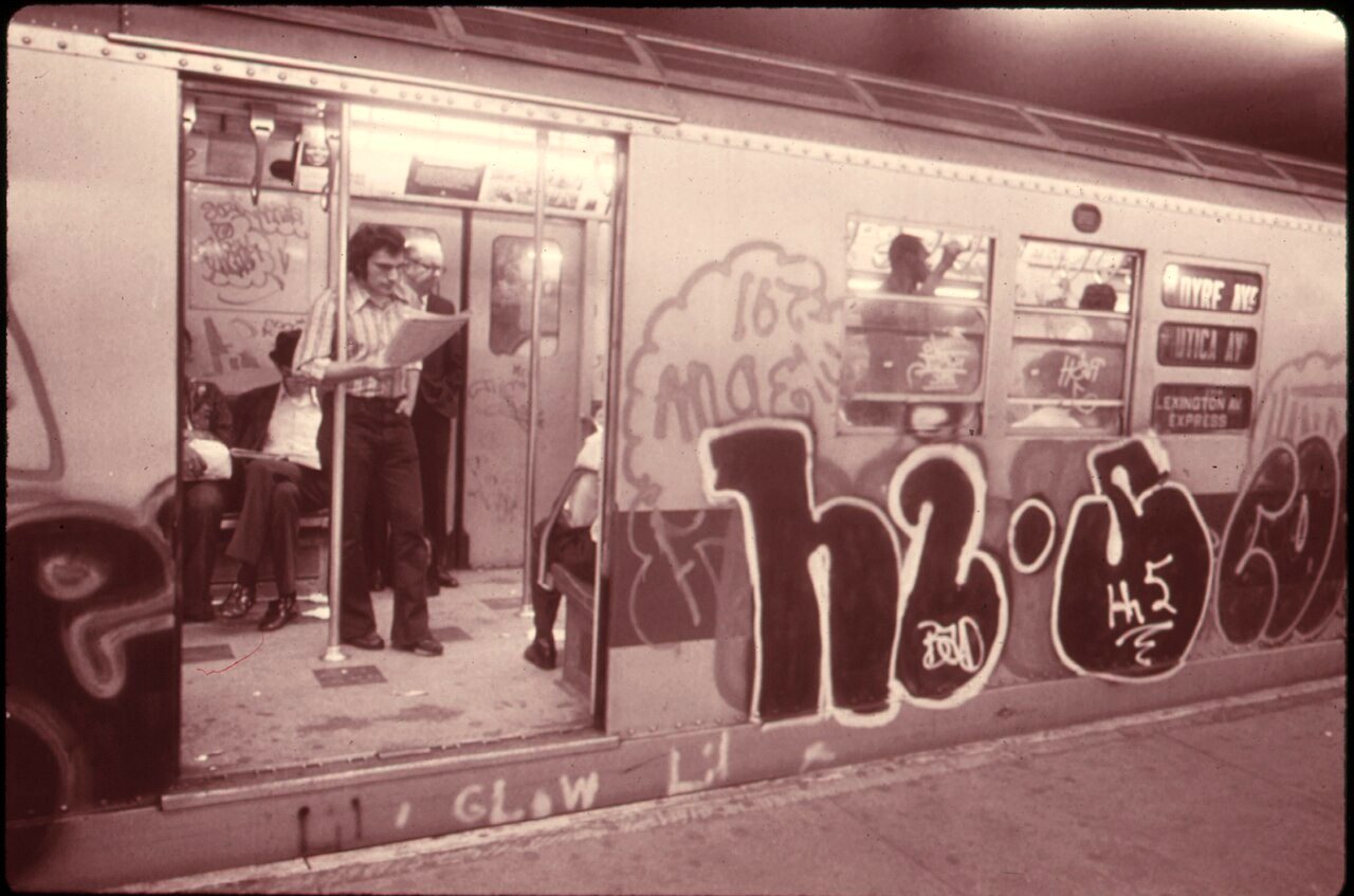 A New York subway car covered in graffiti tags, with passengers visible through its open doors.