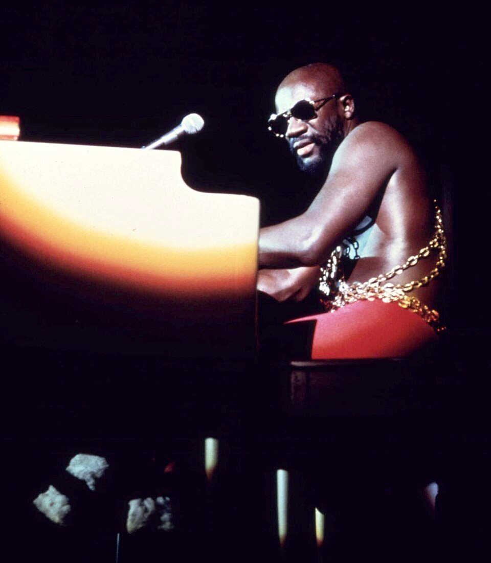 Isaac Hayes in sunglasses and a chain at a piano on a dark stage, a microphone beside him.