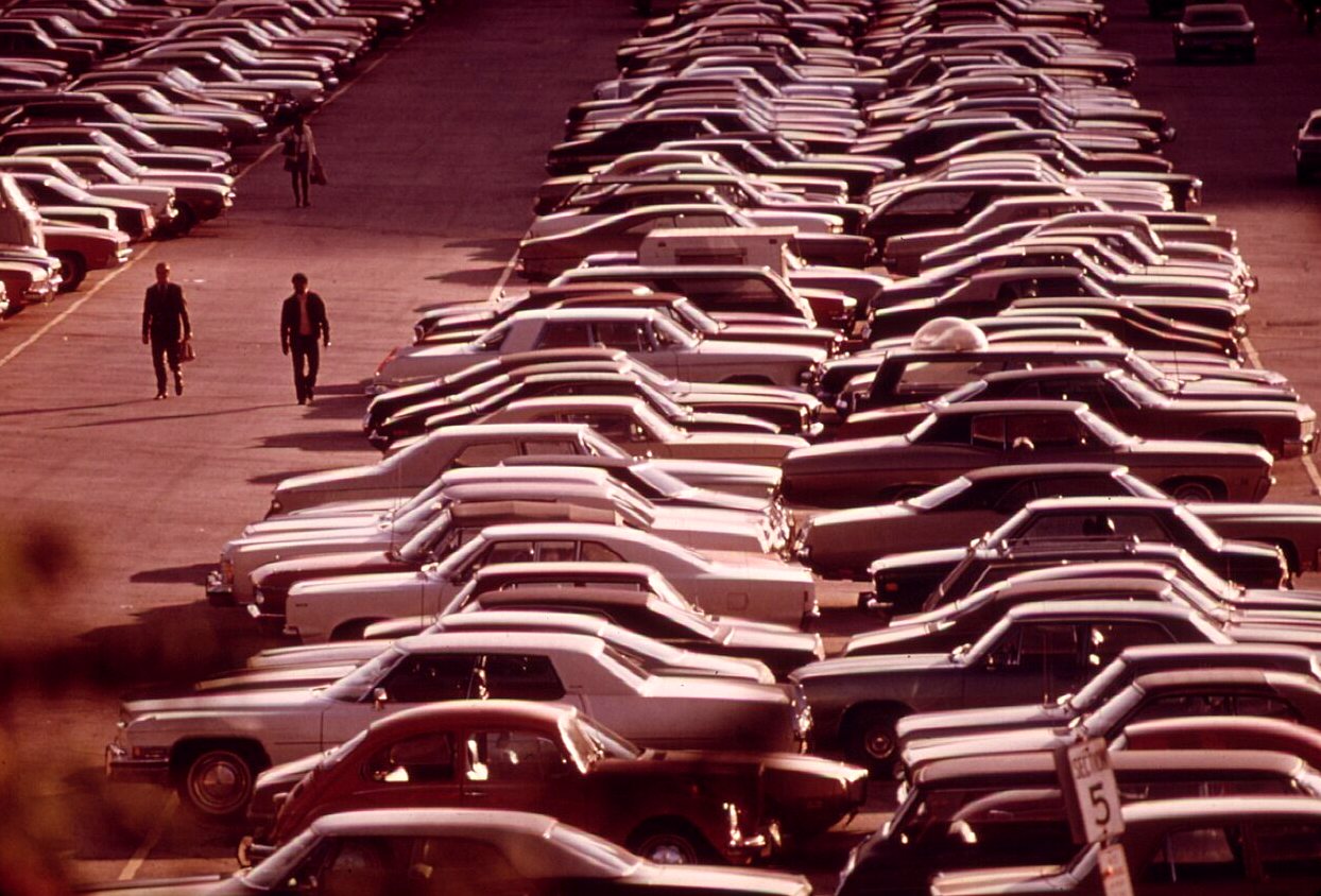 Rows of parked cars seen from above in a huge lot, with two people walking between them.