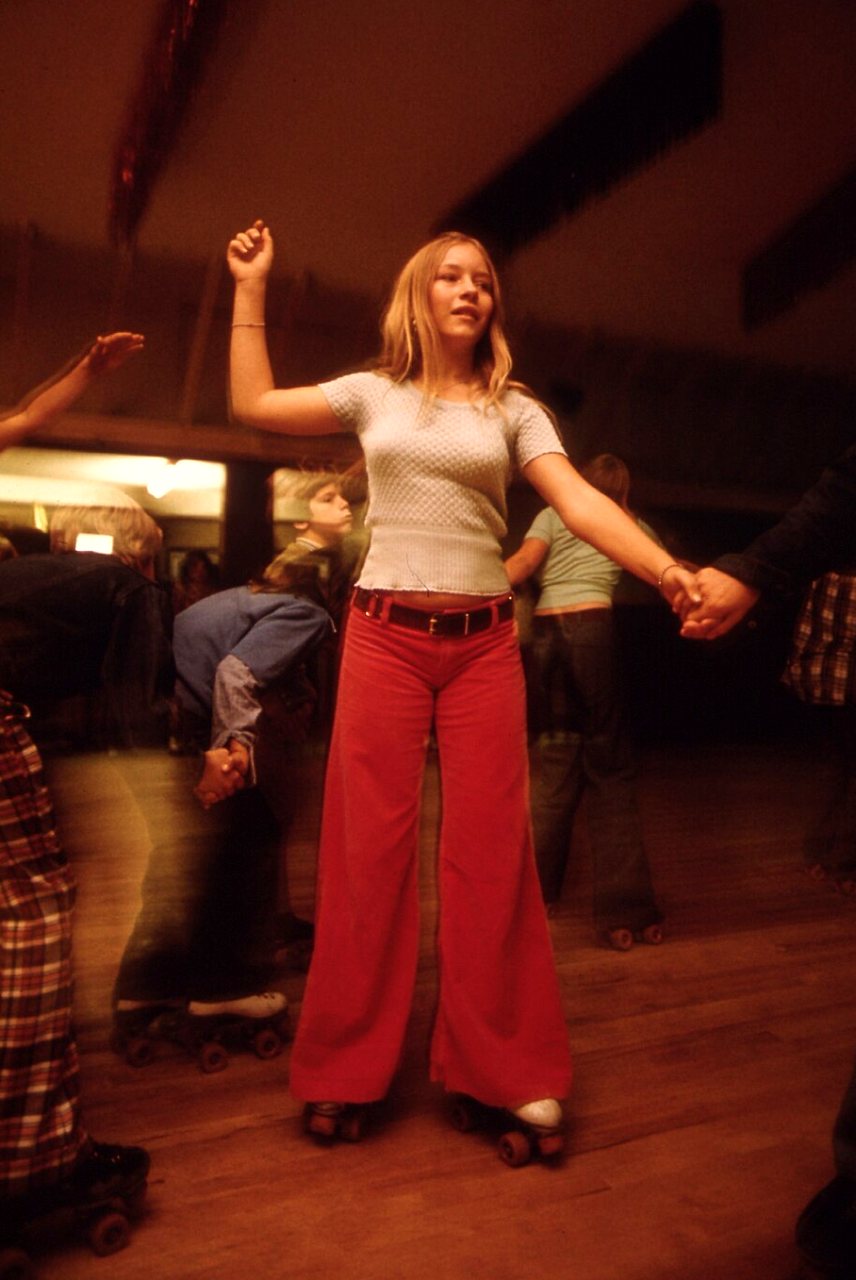 A girl in red flared pants and a tan top roller-skates at a rink, holding hands, with other skaters blurred behind her.