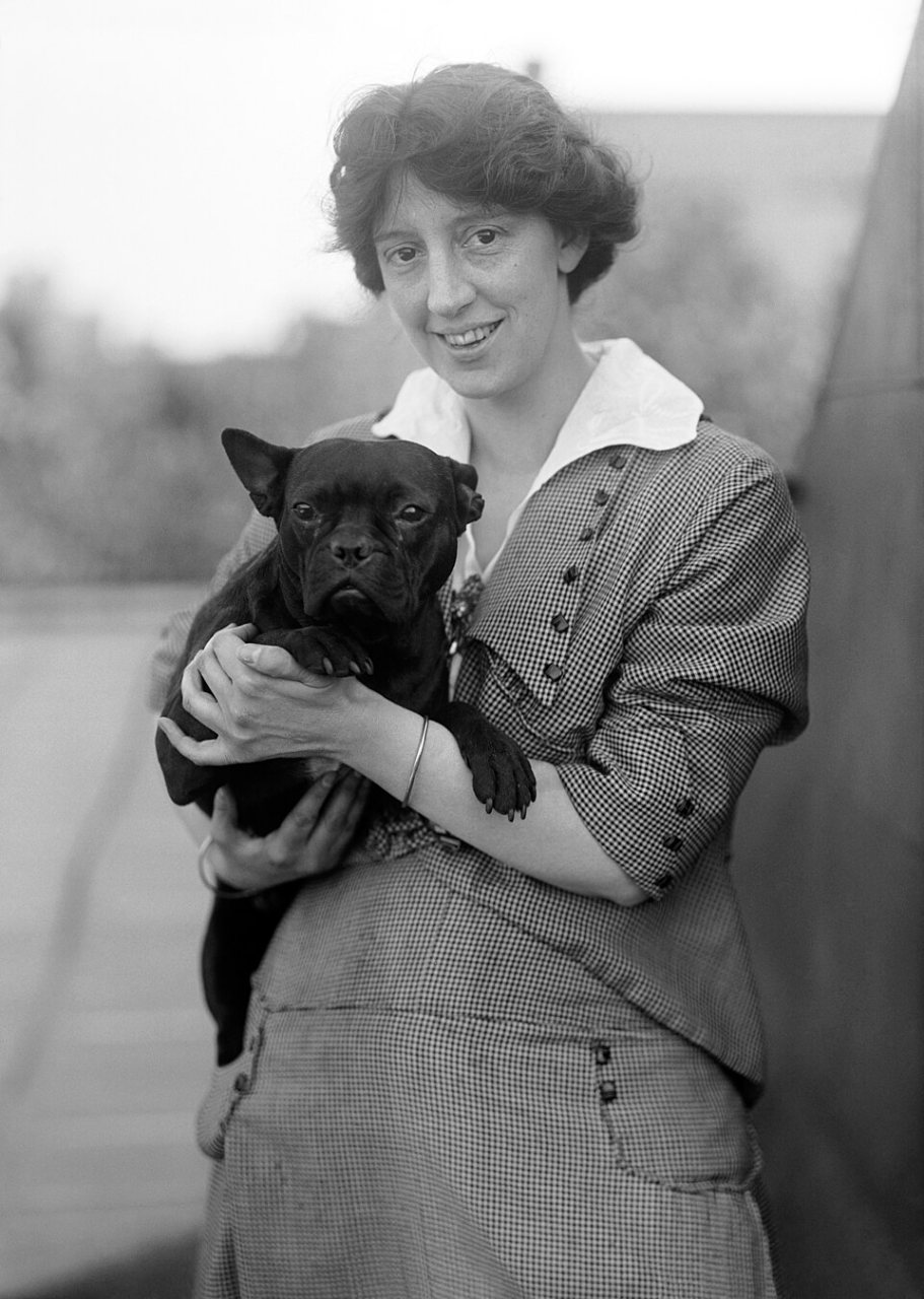 A smiling young woman with pinned-up dark hair, in a white-collared checked dress, holds a small black dog against her chest.