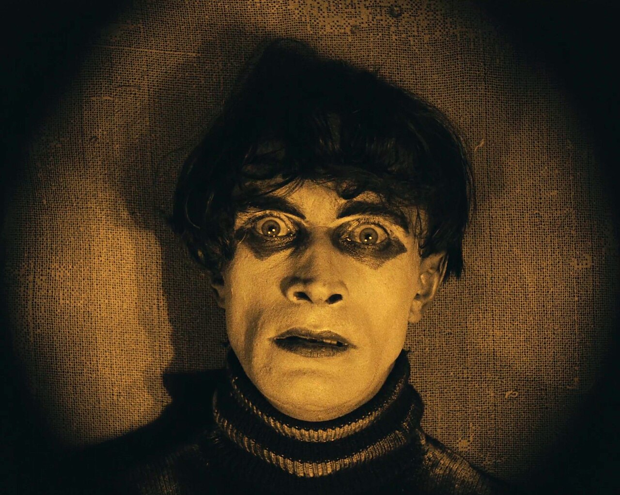A close-up of a young man with a pale face, wide dark-rimmed eyes and black hair falling over his forehead, in a high-necked sweater against a sepia background.