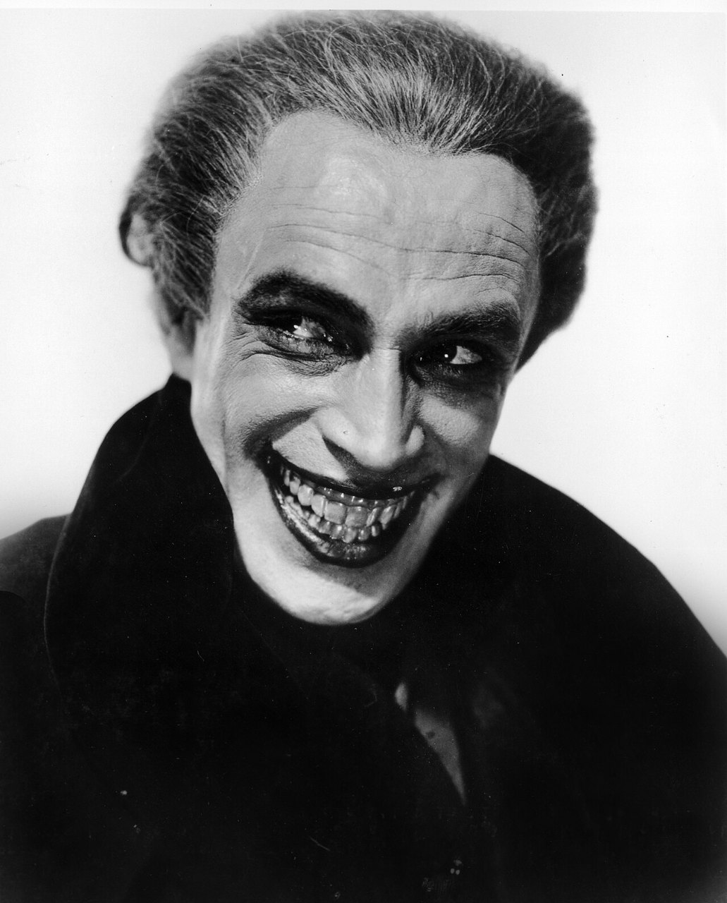 A man with swept-back gray hair and dark-ringed eyes smiles with a fixed, very wide grin full of teeth, in a dark coat against a white background.