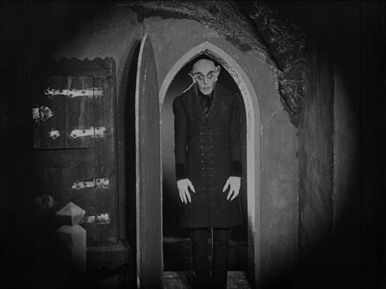 A tall, pale figure in a long dark coat stands stiffly in a narrow Gothic archway, his arms hanging at his sides, in a black-and-white frame that fades to black at the edges.