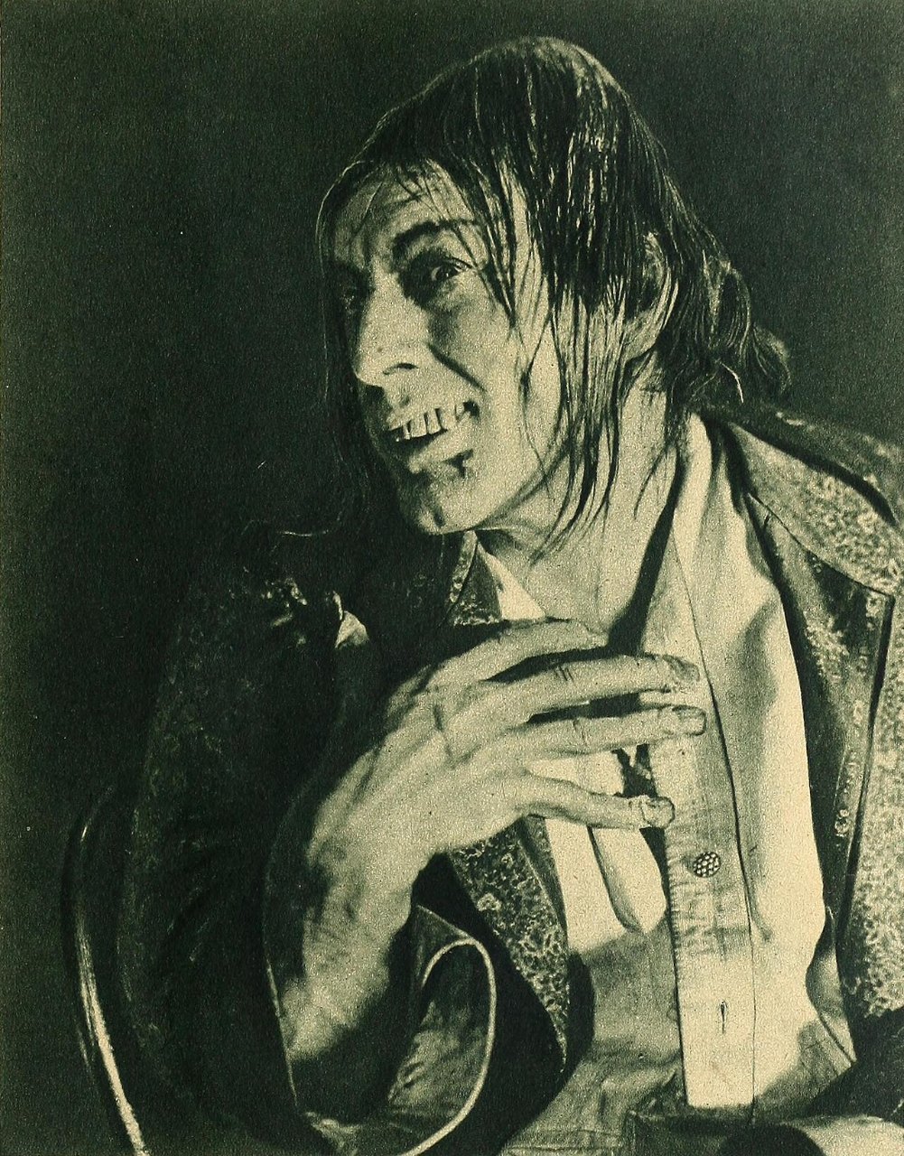 A man with long, damp hair and a sneering grin shows crooked teeth as he leans forward, his thin hands clasped at his chest, in a patterned robe over a white shirt.