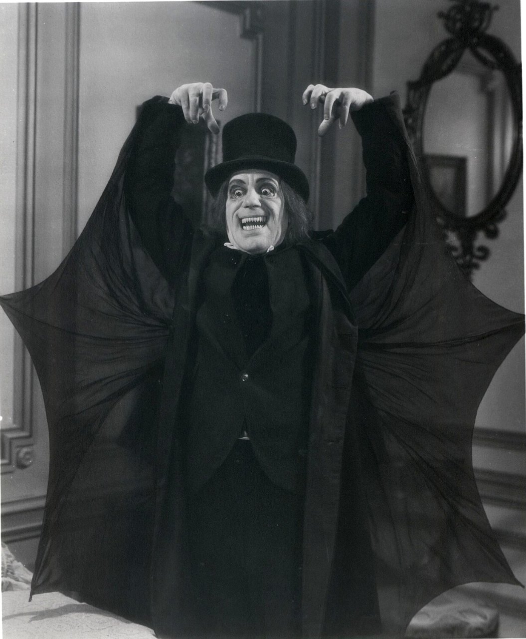 A man in a black top hat and dark suit stands with both hands raised and fingers curled, a sheer black cape stretched between his arms like bat wings, his long hair framing a wide, toothy grin.