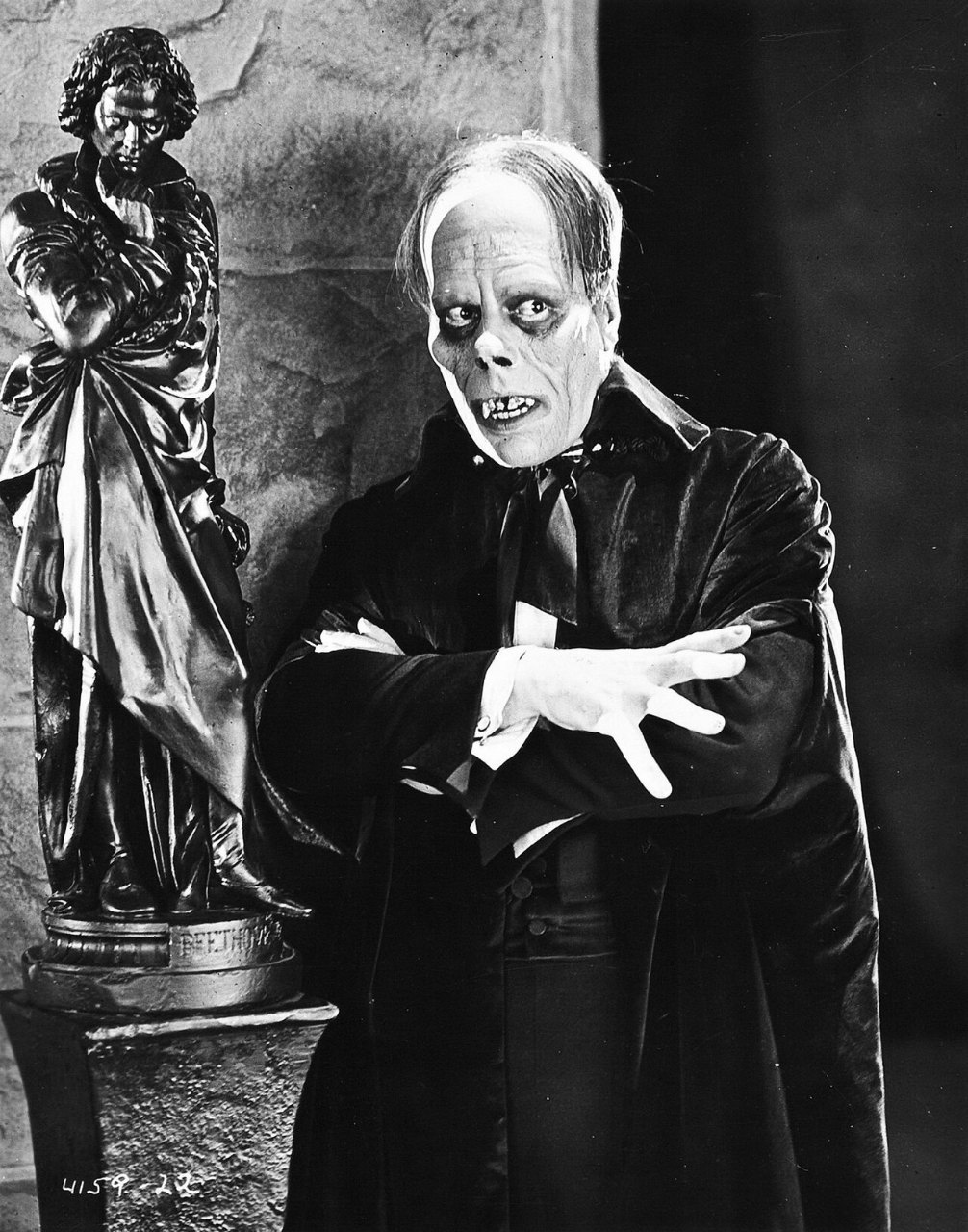 A man with a skull-like face, sunken eyes and bared teeth stands in a black cloak with his arms crossed and fingers curled, beside a statue of a robed figure on a stone pedestal.
