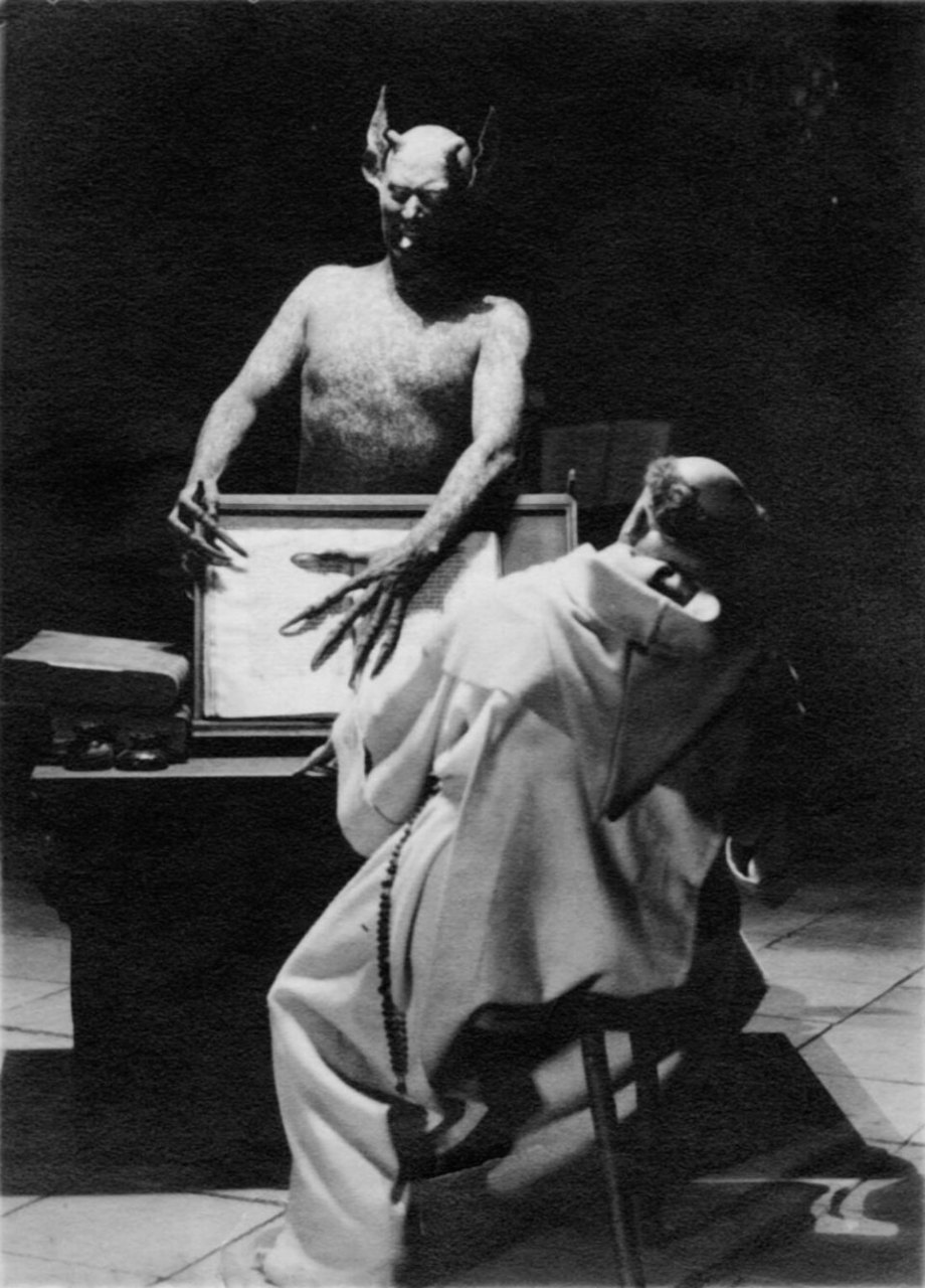 A shirtless, hairy figure with horns, pointed ears and long clawed fingers leans over a framed picture on a table, while a monk in a pale robe sits on a stool with his back to the camera.