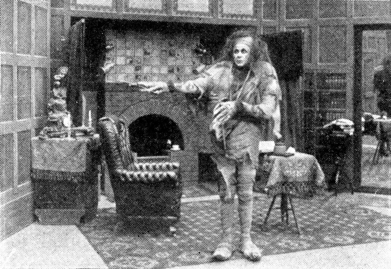A gaunt creature with wild hair and ragged clothes stands with one arm stretched out in a wood-paneled room, a fireplace and an armchair beside him, on a patterned rug.